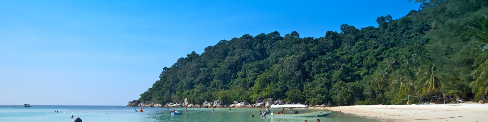 A beach with clear turquoise water, a small boat, and a forested hill in the background.