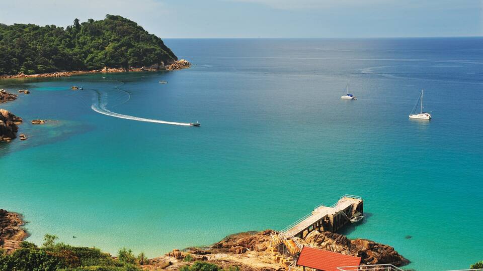 A coastal landscape with a pier, clear turquoise waters, a small boat, and a sailboat.