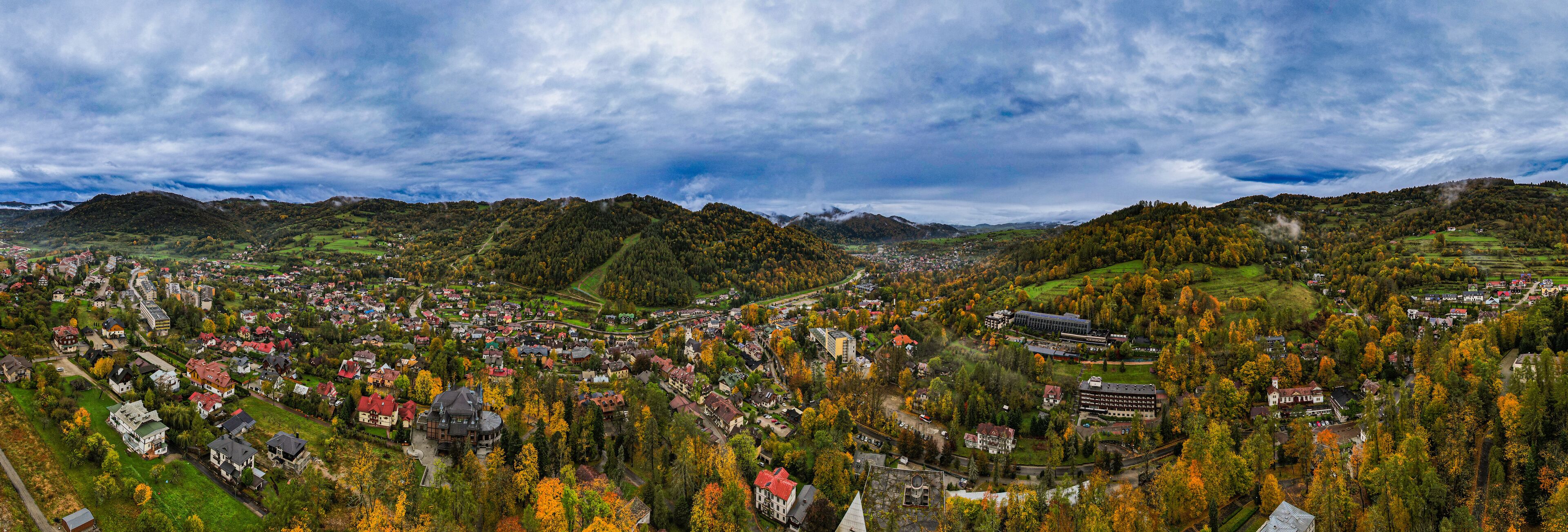 A panoramic view of a town nestled in a valley with autumn foliage, surrounded by hills and mountains.