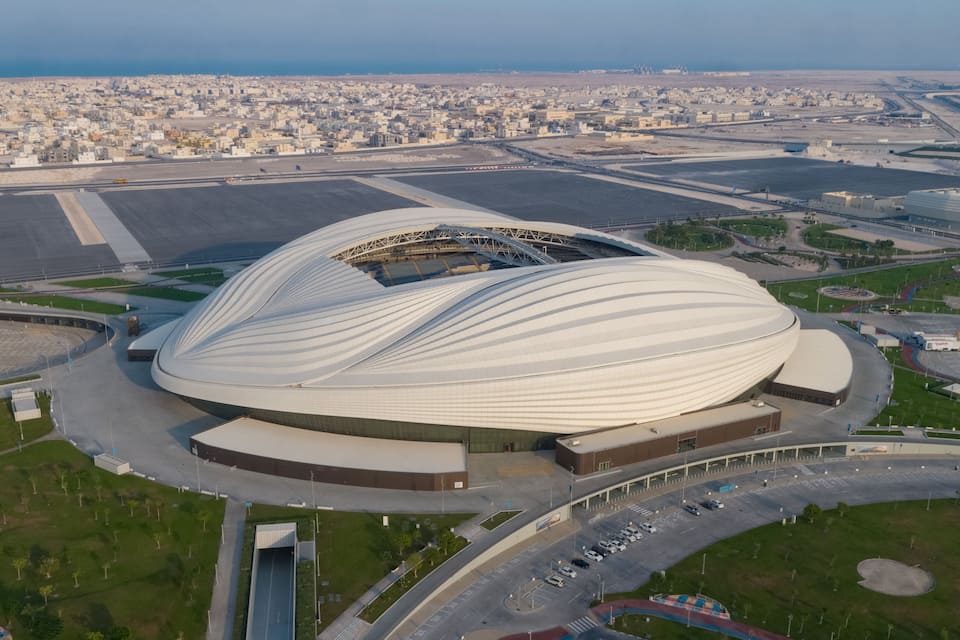 An aerial view of a modern stadium with a unique, curved roof, surrounded by roads and a cityscape in the background.