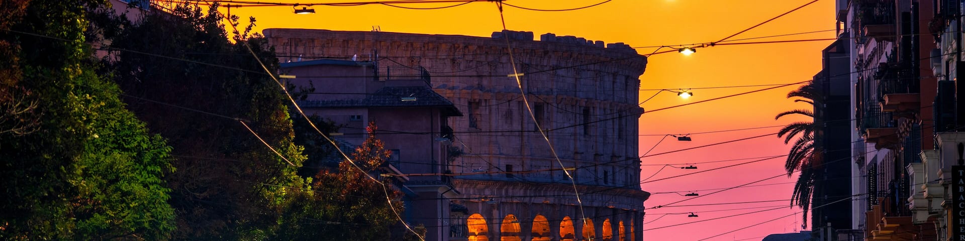A sunset over a historic building with arches, a busy street with cars, and power lines.