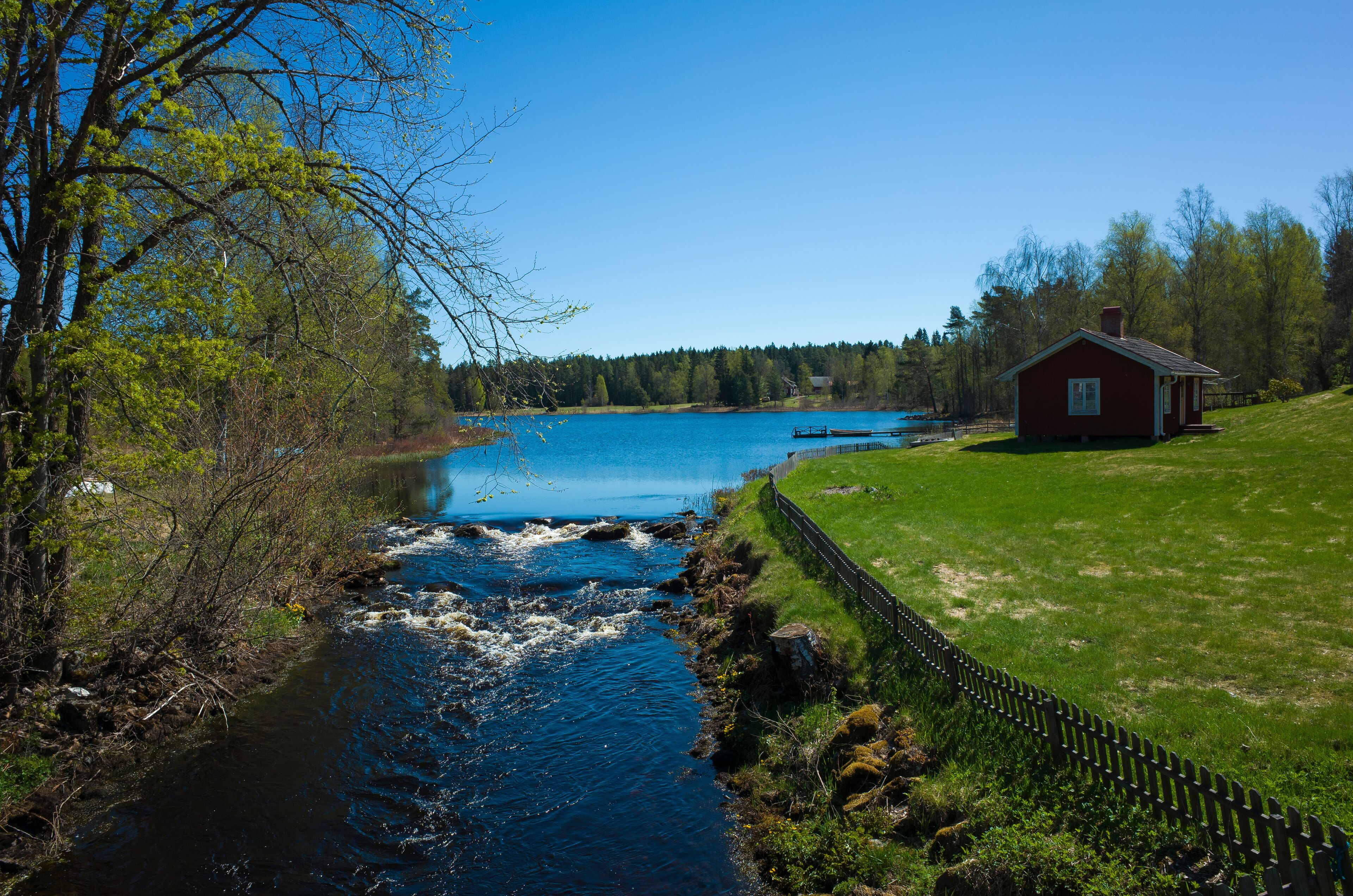 A river flowing through a grassy area with a wooden fence and a red house in the background.