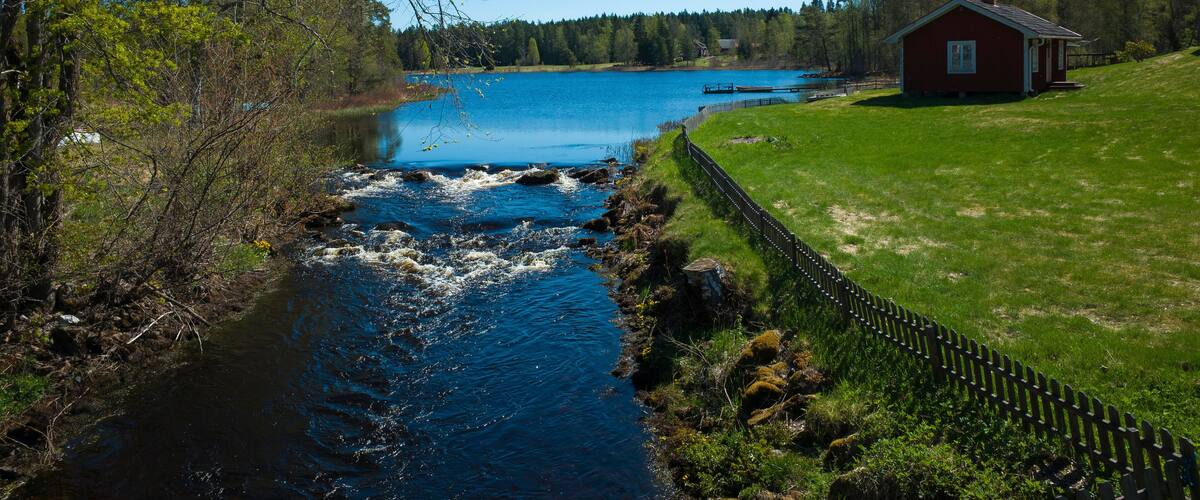 A river flowing through a grassy area with a wooden fence and a red house in the background.