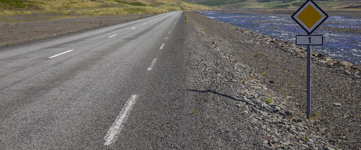 A road with a speed limit sign of 90 km/h, a diamond-shaped warning sign, and a mountainous landscape with a river.