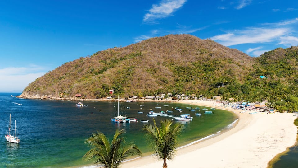A coastal beach with boats, a sailboat, and a mountainous backdrop.