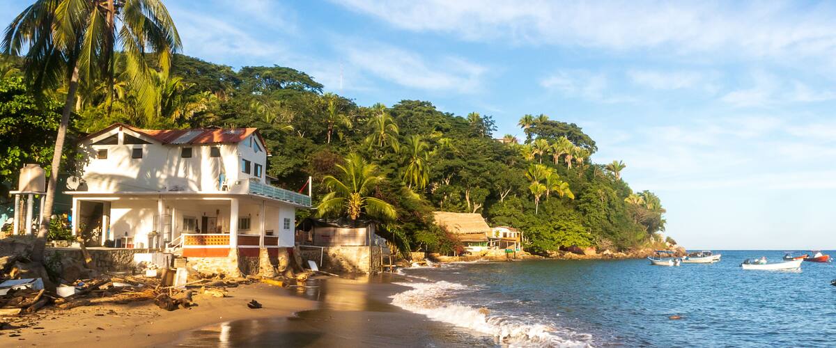A beach with a white house, palm trees, and boats in the water.