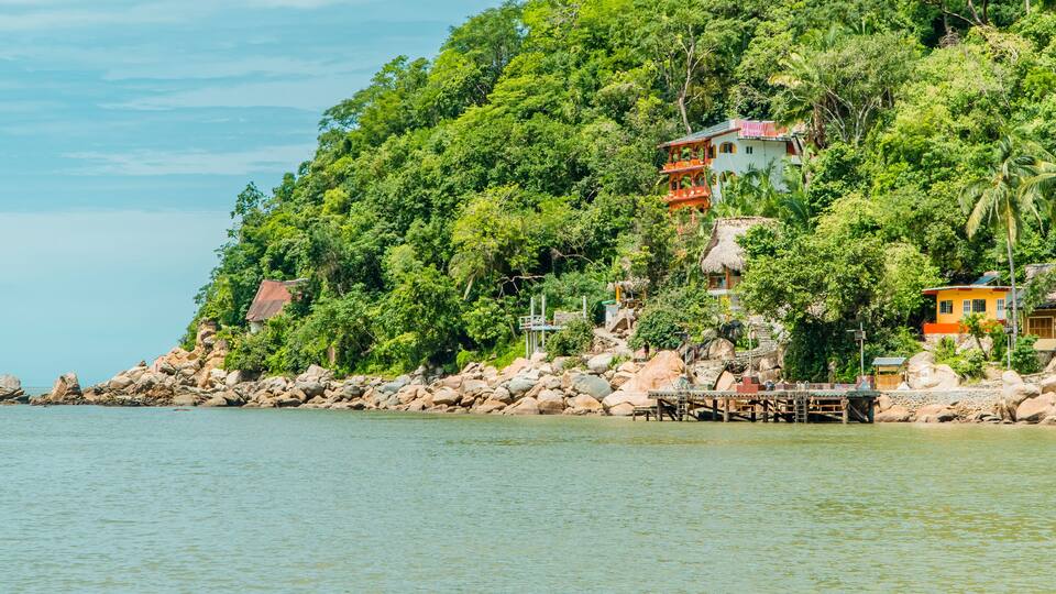 A coastal area with a rocky shoreline, a thatched-roof house, and a colorful building on a hillside.