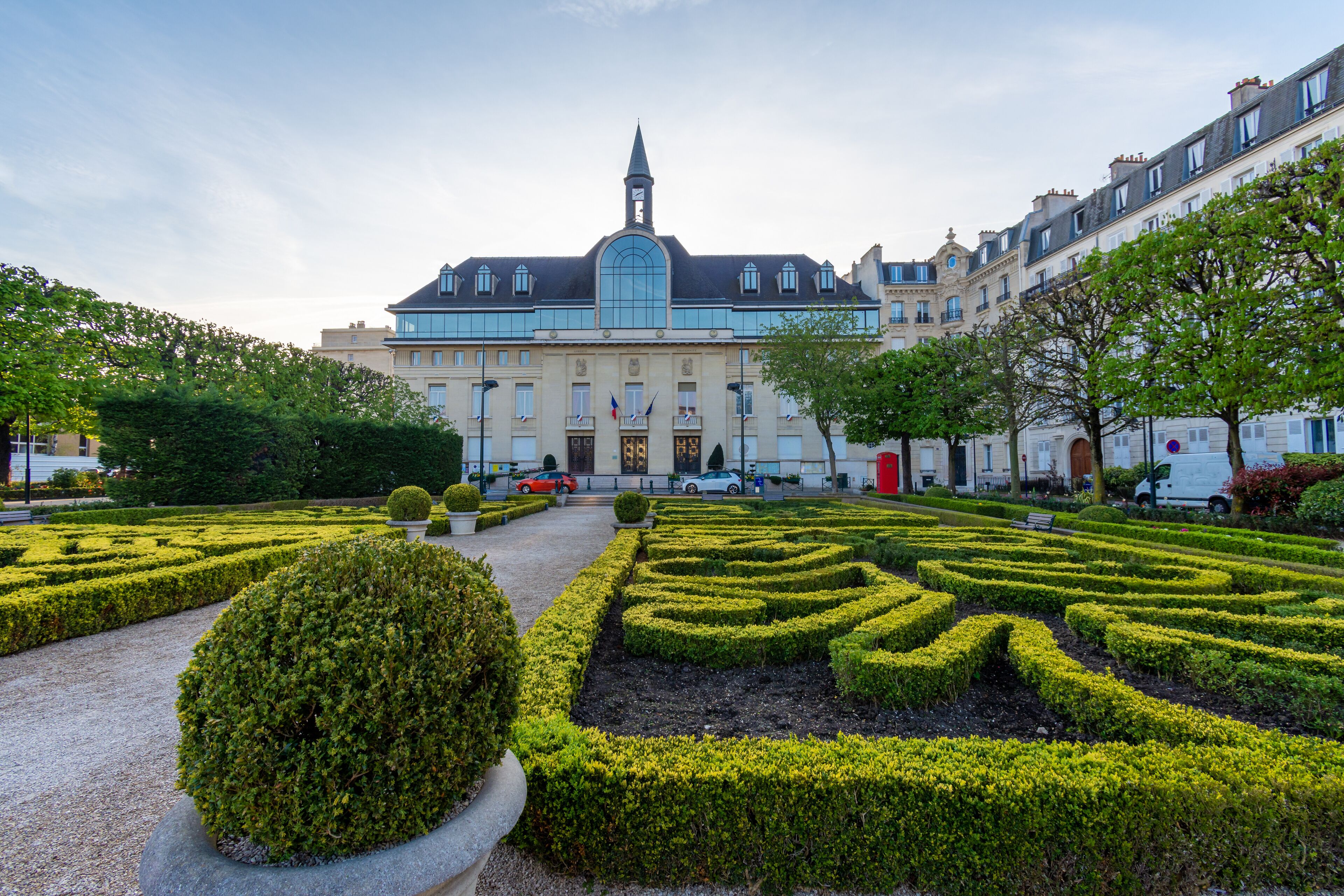 A formal garden with geometrically trimmed hedges and a large building with a clock tower in the background.