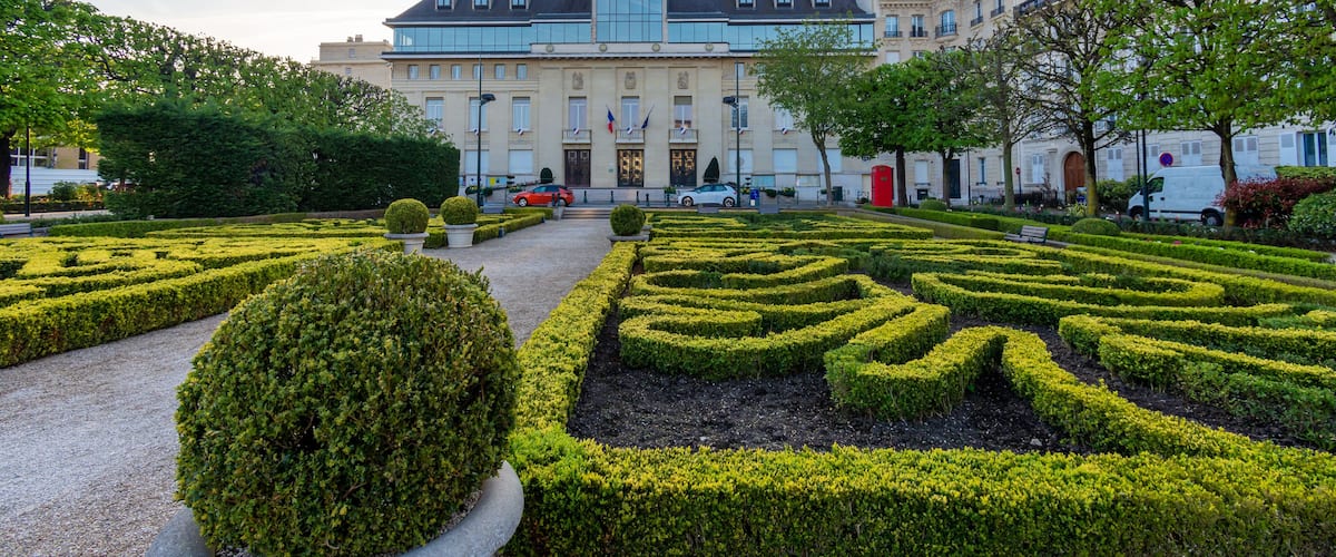 A formal garden with geometrically trimmed hedges and a large building with a clock tower in the background.