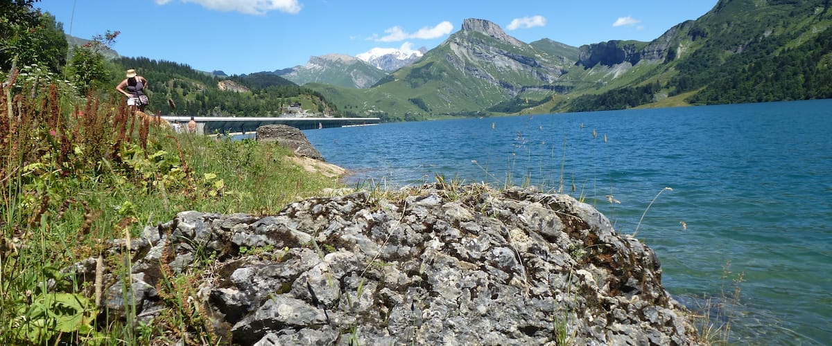 A rocky shoreline with a person standing, a bridge in the background, and mountains in the distance.