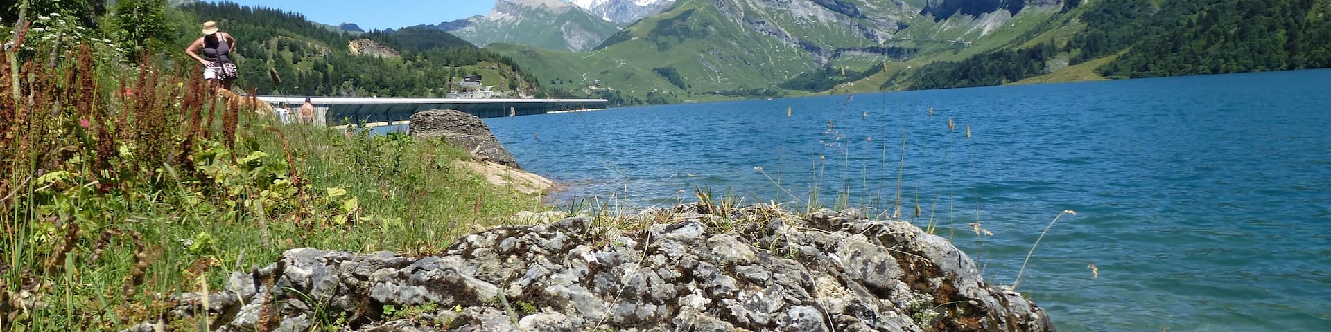 A rocky shoreline with a person standing, a bridge in the background, and mountains in the distance.