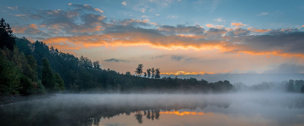 A misty lake with a forested shoreline and a sunset sky.