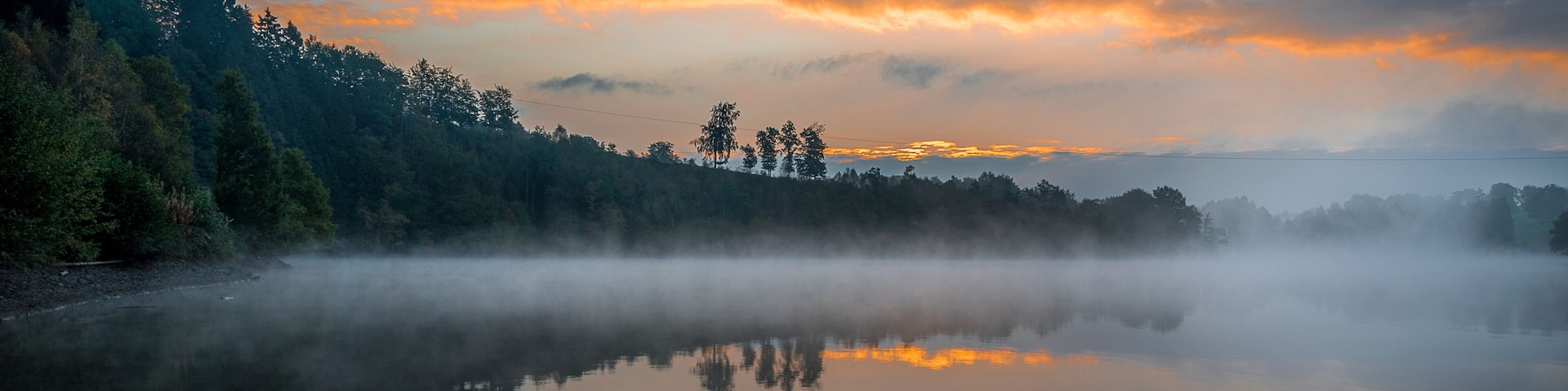A misty lake with a forested shoreline and a sunset sky.