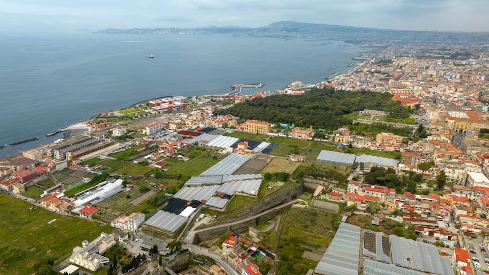 An aerial view of a coastal city with a mix of residential and industrial areas, including greenhouses and a port with ships.