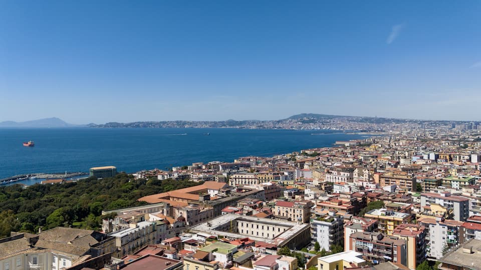 A coastal cityscape with a view of the sea, a ship, and a distant mountain.