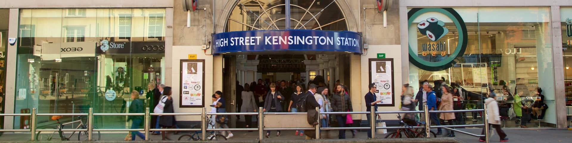 A busy street with a prominent High Street Kensington Station sign, a glass-fronted store, and a sushi restaurant.