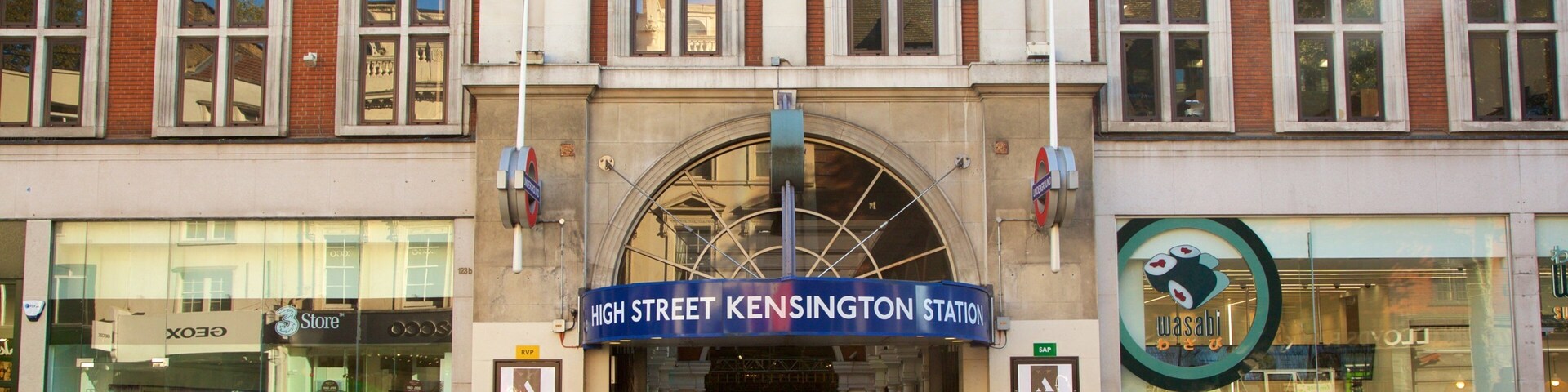 Una calle concurrida con un llamativo letrero de la estación High Street Kensington, una tienda con fachada de vidrio y un restaurante de sushi.