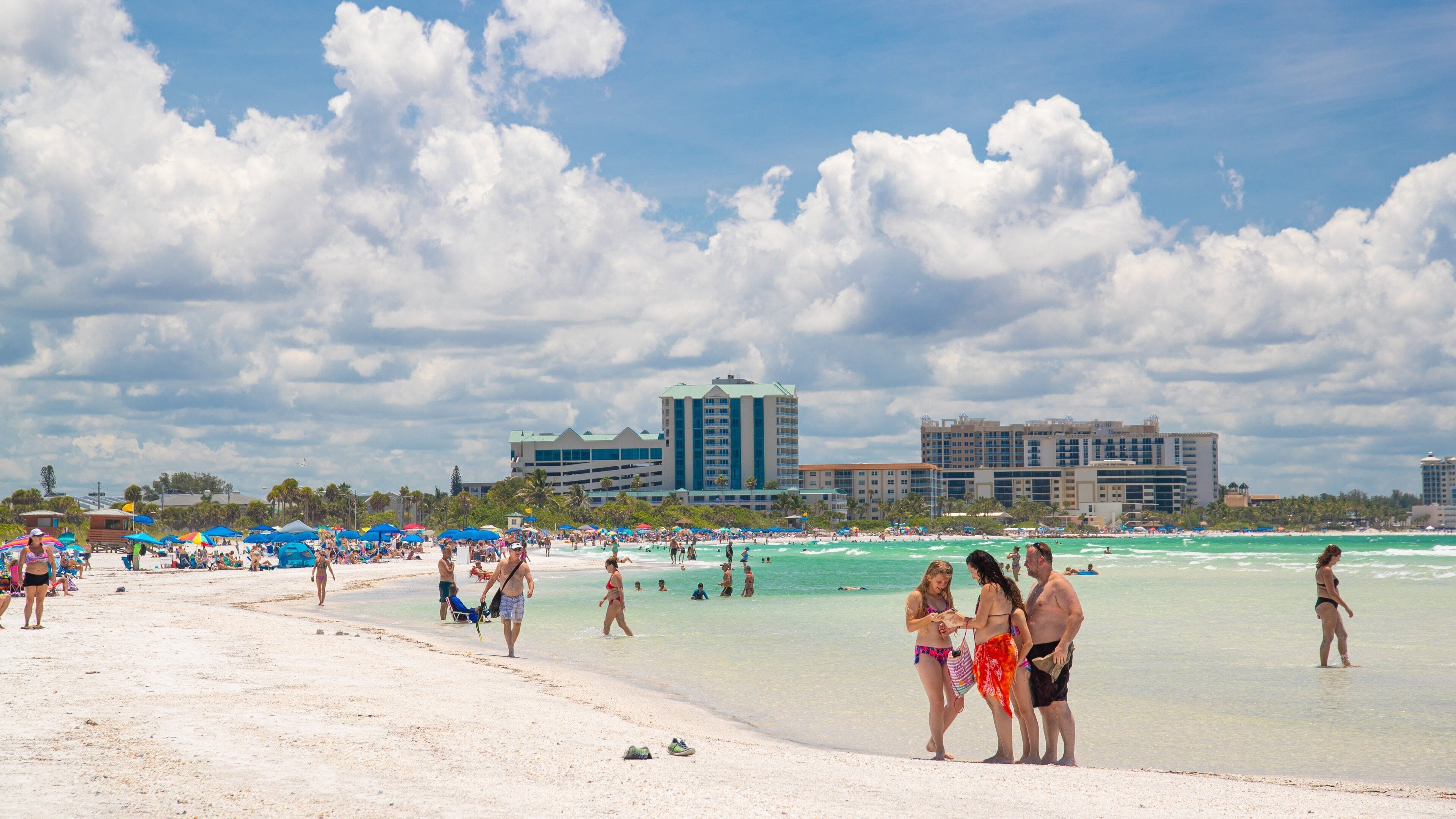A crowded beach with people sunbathing and swimming, a clear sky, and hotel buildings in the background.