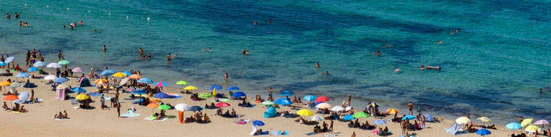 Una playa concurrida, gente tomando el sol y nadando, con sombrillas de colores.