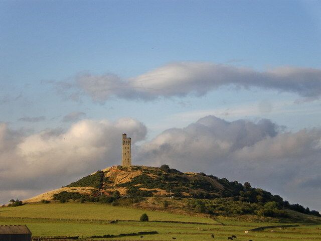Uma torre numa colina com campos verdes e nuvens no céu.