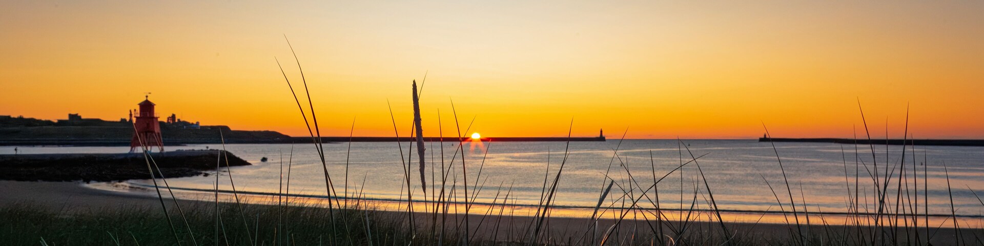 Un atardecer sobre un mar tranquilo con un faro y un primer plano con hierba.