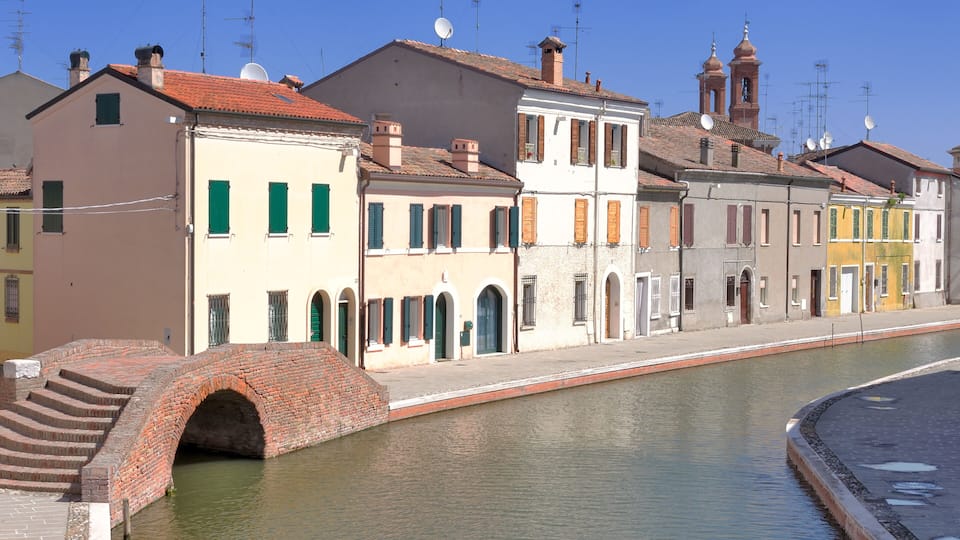 A canal with a brick bridge, historic buildings, and a church with two towers.