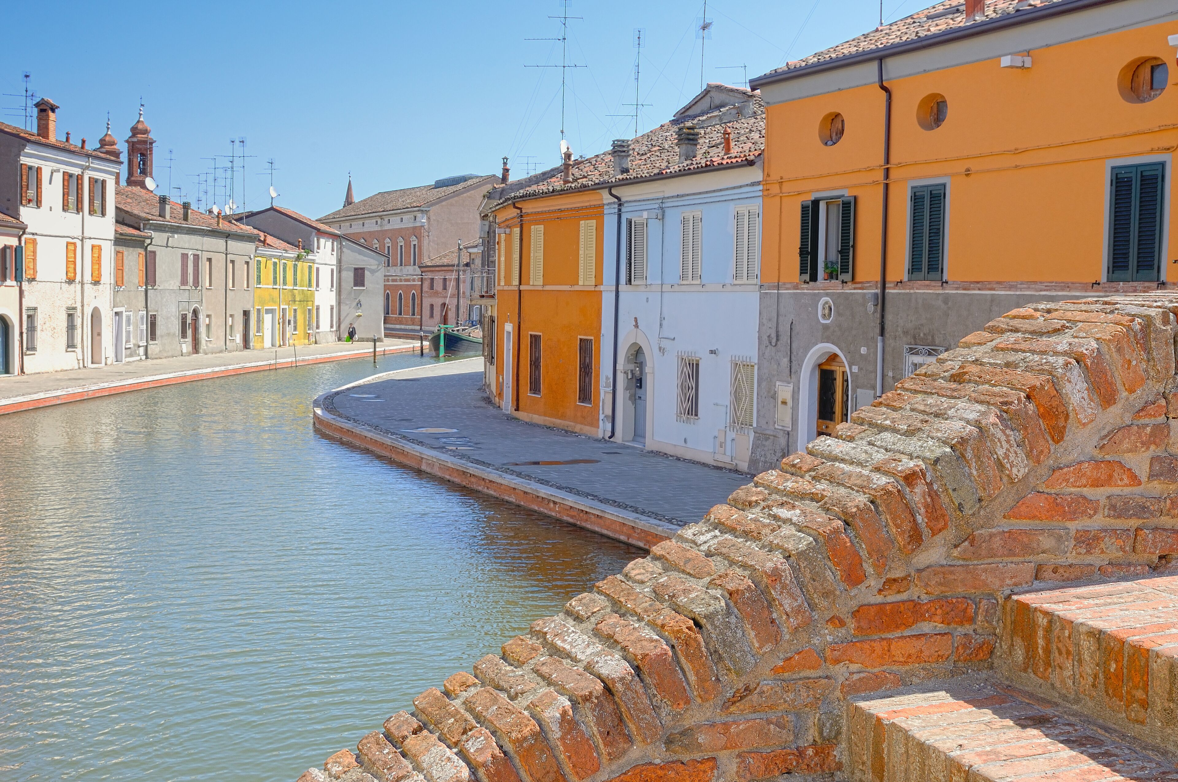 A canal with buildings on either side, a stone bridge, and a clear blue sky.