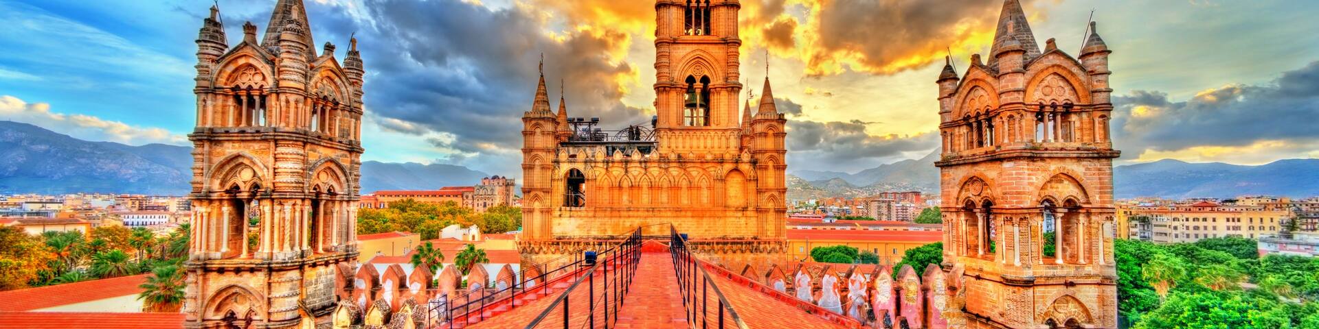 A historic cathedral with twin towers and a central bell tower, surrounded by a red-roofed plaza and palm trees.