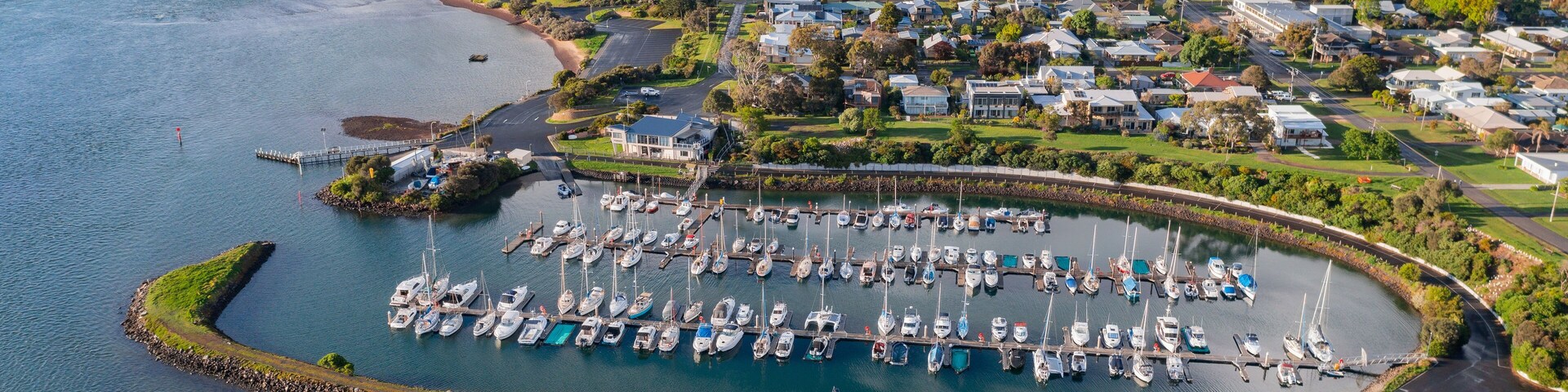 Una marina con numerosos barcos amarrados, un puente al fondo y un pueblo costero con casas y vegetación.