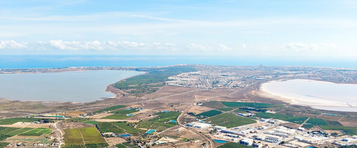 An aerial view of a coastal city with a large body of water, green agricultural fields, and industrial areas.