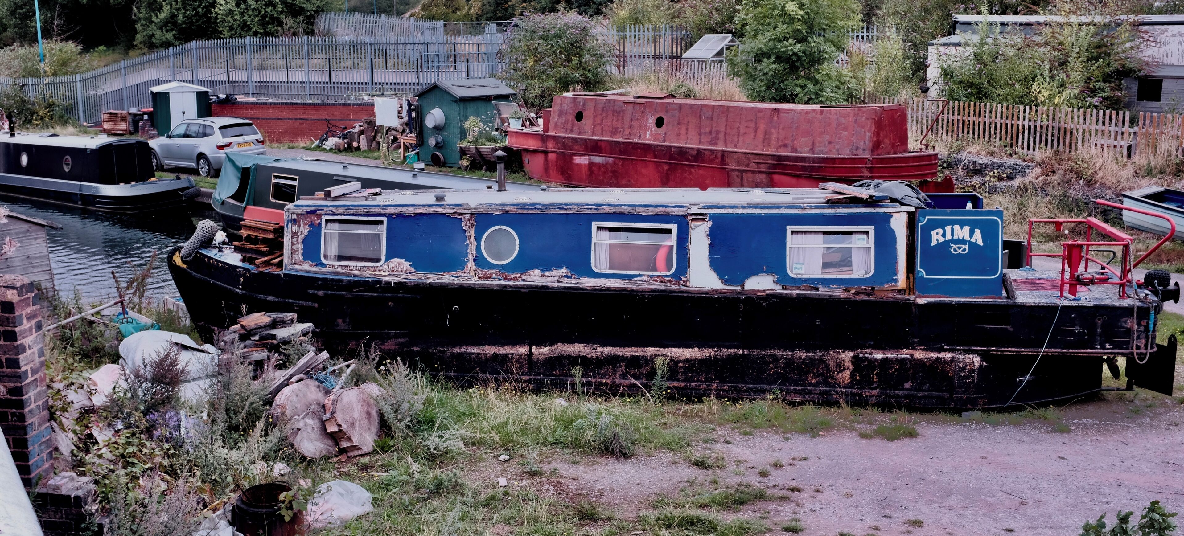 A derelict boat with the name "RIMA" on its side, surrounded by overgrown vegetation and other abandoned structures.