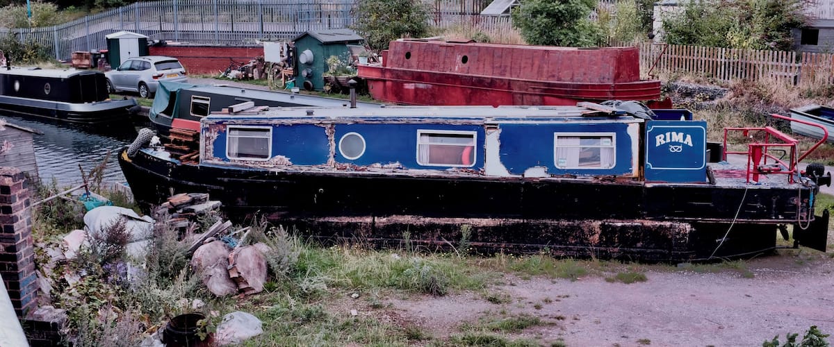 A derelict boat with the name "RIMA" on its side, surrounded by overgrown vegetation and other abandoned structures.