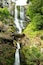 Ein Wasserfall stürzt über eine felsige Klippe, umgeben von üppiger Vegetation.