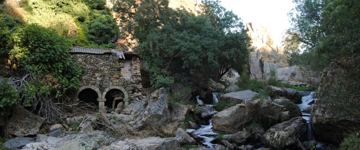 A stone bridge with three arches over a rocky stream, surrounded by lush greenery and a mountainous backdrop.