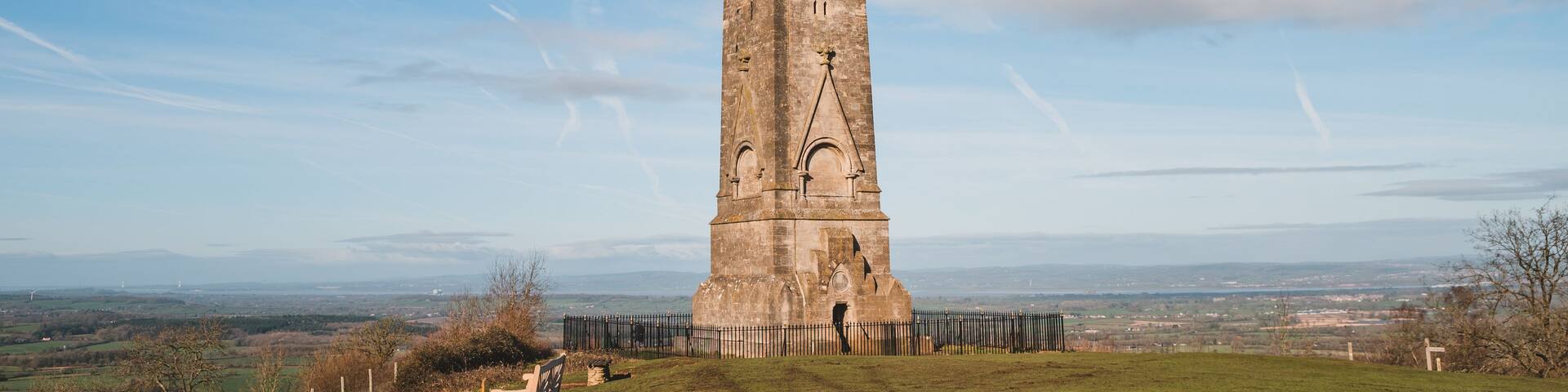 A tall, historic stone tower with a cross on top, surrounded by a grassy field and a clear blue sky.