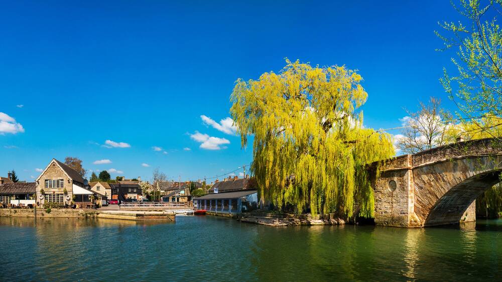 A stone bridge over a river, a weeping willow tree, and traditional buildings along the riverbank.