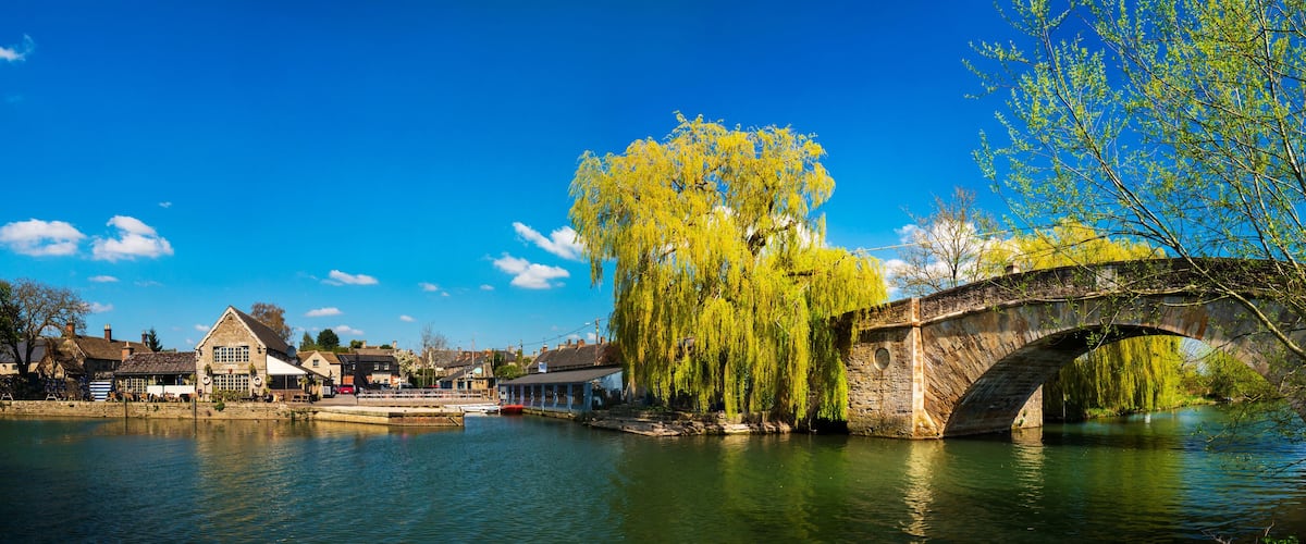 A stone bridge over a river, a weeping willow tree, and traditional buildings along the riverbank.