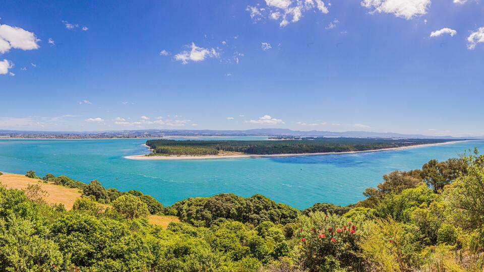 A coastal landscape with a clear blue sea, a sandy beach, and dense green vegetation.