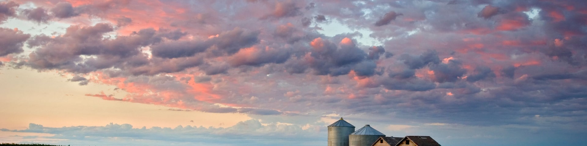 Paisaje rural con un campo de hierba alta, dos silos de grano y un granero de madera bajo un cielo colorido.