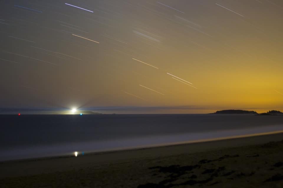 Una playa al atardecer con estelas de estrellas en el cielo y un mar en calma.