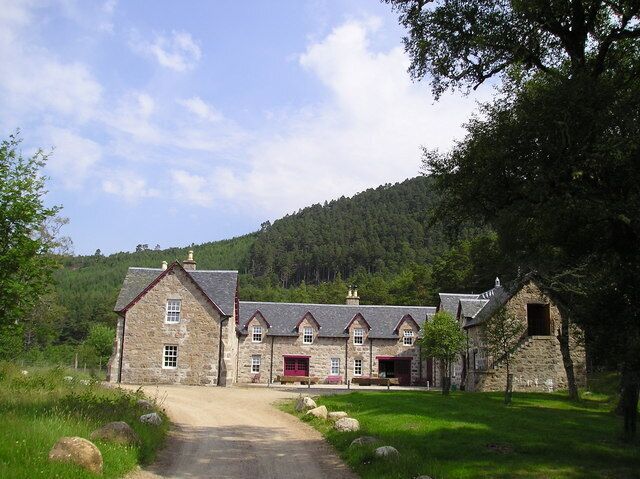 A stone building with a red door, surrounded by a grassy area and trees, with a hill in the background.