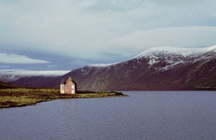A small pink church on a grassy island in a lake, surrounded by mountains.