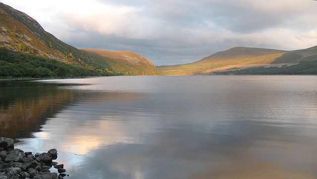 A calm lake with rocky shores and hills in the background.