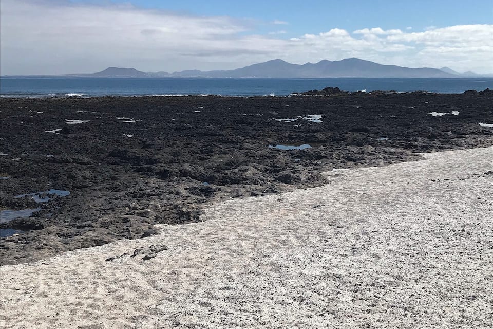 Une côte rocheuse, sous un ciel dégagé, avec des montagnes qui se dessinent à l’horizon.