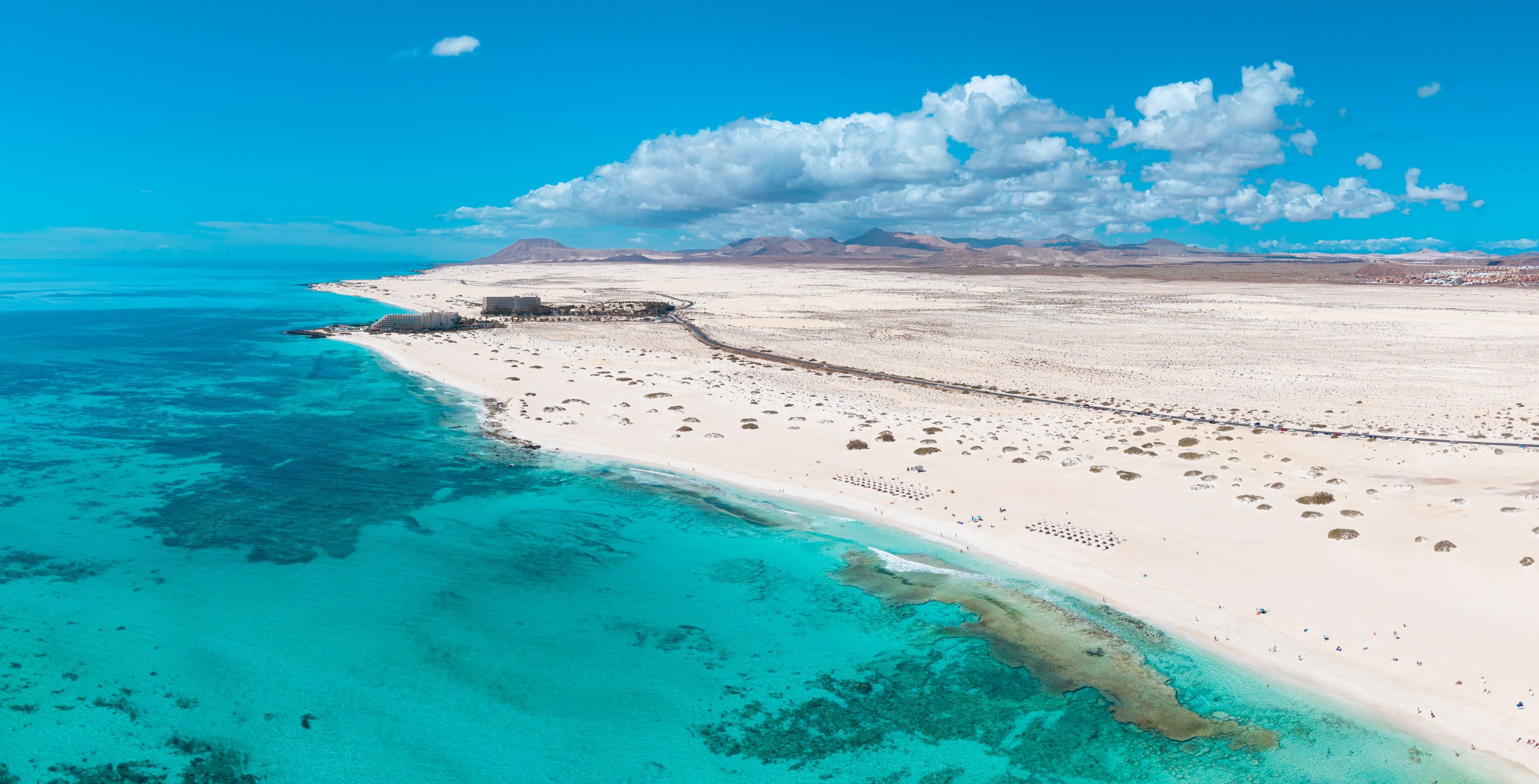 A coastal landscape with a sandy beach, clear turquoise waters, and distant mountains under a blue sky.