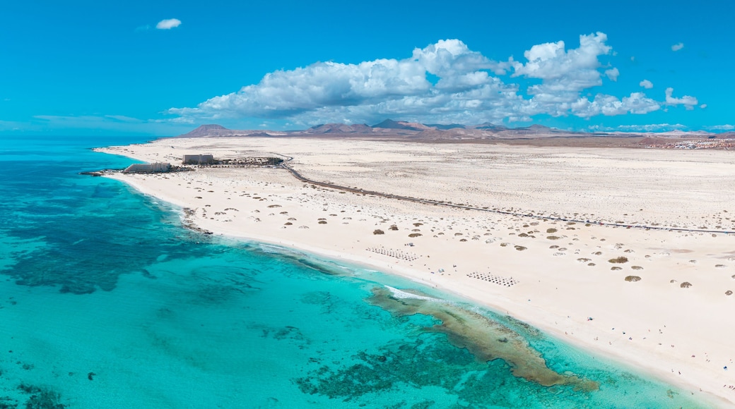 A coastal landscape with a sandy beach, clear turquoise waters, and distant mountains under a blue sky.