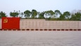 Una plaza pavimentada con un muro rojo que muestra un emblema de hoz y martillo, y un muro de piedra con texto grabado.