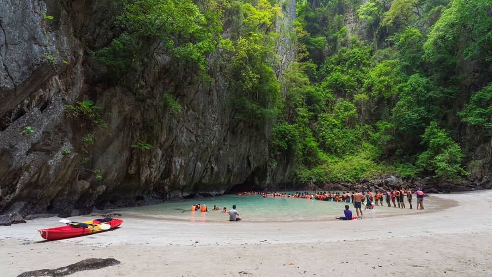A beach with kayaks, a group of people in the water, and a rocky cliff with greenery.