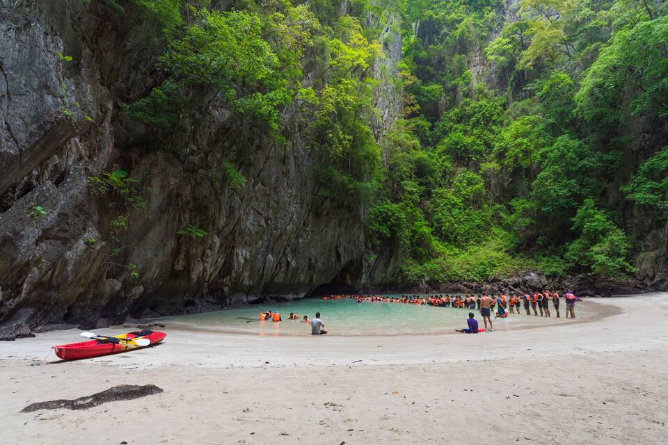 Una playa con kayaks, un grupo de personas en el agua y un acantilado rocoso con vegetación.