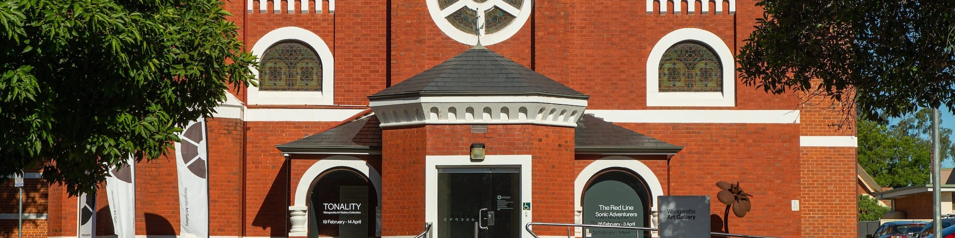A red brick building with a large circular stained glass window, a white entrance, and a small bell tower.